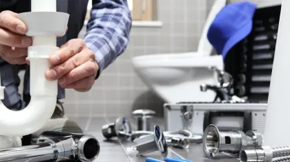 Plumber assembling white PVC pipe under a sink amid plumbing tools and parts in a tiled bathroom.