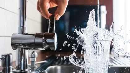 Hand turning on modern kitchen faucet with water splashing out of a glass in stainless steel sink.