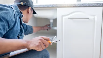 Plumber inspecting and repairing white kitchen cabinet plumbing with adjustable wrench in hand.