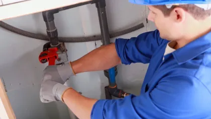 Plumber in blue uniform and hard hat repairing pipe under sink with red pipe wrench and gloves