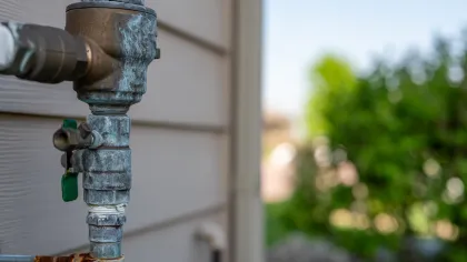 Close-up of a rusty and weathered water spigot attached to an exterior wall with green foliage in background