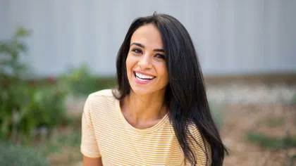 Smiling woman with long dark hair wearing a yellow striped t-shirt outdoors with blurred background.