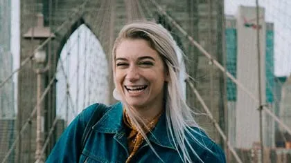 Smiling young woman with blonde hair and denim jacket on Brooklyn Bridge in New York City.