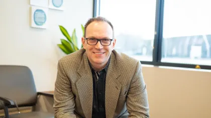 Smiling man wearing glasses and a blazer sitting in a modern office with natural light from a window.