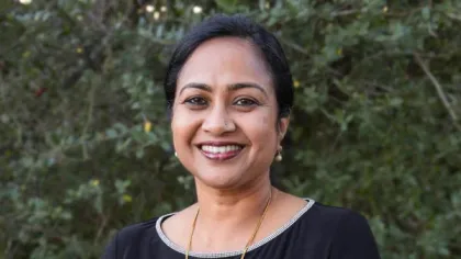 Smiling woman with dark hair in a black top standing outdoors with green foliage background.