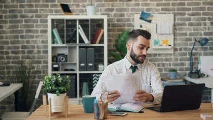 Man working at desk with laptop and documents in a modern office with brick wall and shelf.