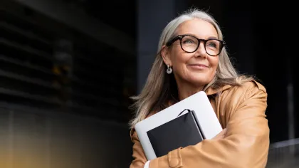 Smiling mature woman in glasses and tan coat hugging a laptop and notebook outdoors with confident expression