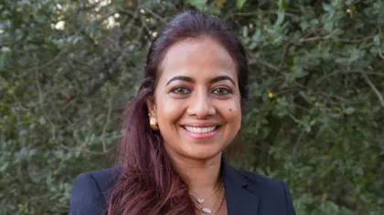 Smiling woman with long dark hair wearing a black blazer and gold jewelry outdoors with green foliage background
