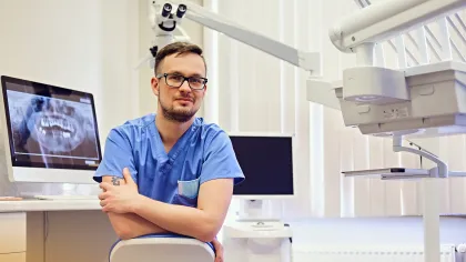 Male dentist in blue scrubs sitting in dental office with X-ray and dental equipment in the background.