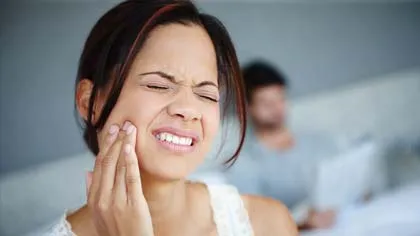 Woman with dark hair touching her cheek, showing facial pain or discomfort indoors with a blurred person in background.