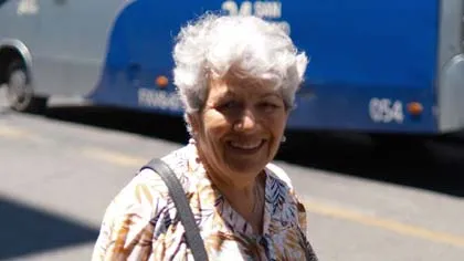 Smiling elderly woman with white hair wearing patterned blouse and shoulder bag outdoors near a blue bus.