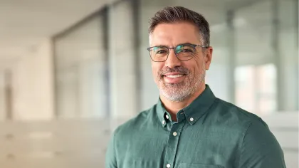 Smiling middle-aged man with glasses wearing green shirt holding a tablet in a modern office space.