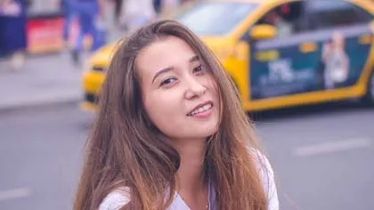 Smiling young woman with long hair posing on city street with yellow taxi in background.