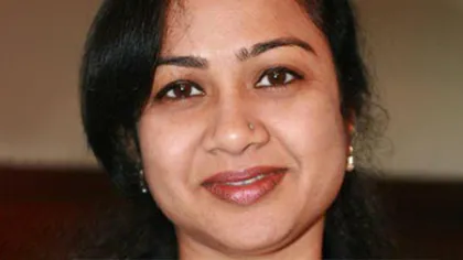 Close-up portrait of a smiling woman with black hair, brown eyes, and a nose piercing indoors.