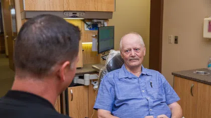 Older man in blue shirt sitting in a medical office talking to a healthcare professional.