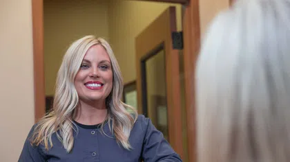 Smiling blonde woman in gray scrub top stands behind reception desk in a professional office environment.