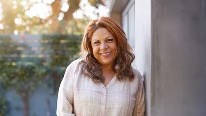 Smiling young woman with long hair wearing a blue shirt, standing in front of green shutters.