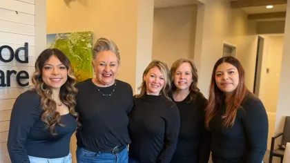 Group of five women standing indoors smiling, dressed in black tops and jeans, in a bright office setting.