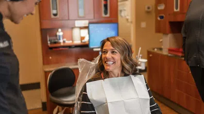 Woman smiling in dental chair with dental bib talking to a standing dental professional in a clinic.
