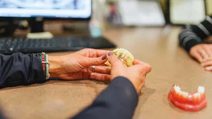 Hands holding a dental model with braces on a desk, computer and another dental model in the background.