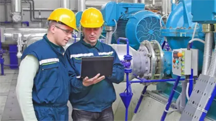 Two engineers in yellow helmets checking a laptop inside an industrial plant with machinery and pipes.
