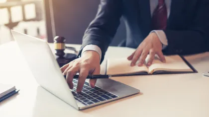 Businessman in suit using laptop and taking notes with a pen in office setting with gavel nearby.
