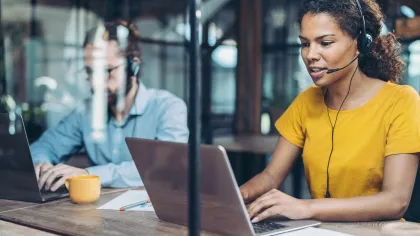 Woman in yellow shirt wearing headset working on laptop in office with male colleague in background.