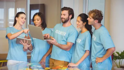 Group of volunteers in blue shirts collaborating using laptop and tablet around a table with colorful notes.
