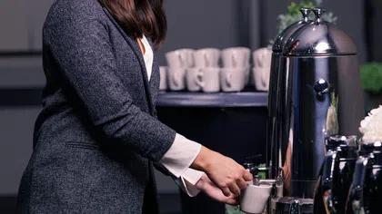 Woman in gray blazer pouring coffee from a large shiny urn at a coffee station with cups in background.