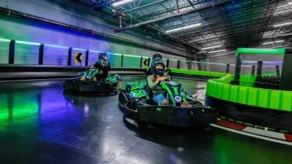 Two racers in go-karts speed around a vibrant indoor track illuminated with colorful lights.