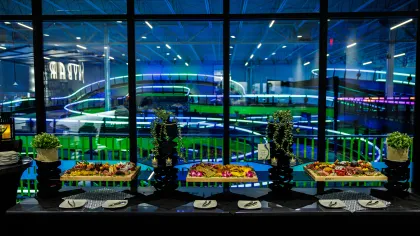 Catering table with appetizers and plants in front of large window showing indoor go-kart track with neon lights.