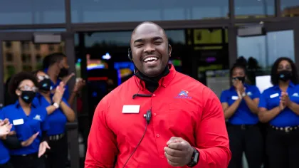 Smiling employee in red shirt welcomes guests outside, with team members in blue shirts clapping.