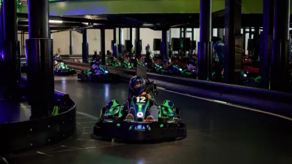 Indoor go-karting track with racers in helmets driving green and black karts under dim lighting.