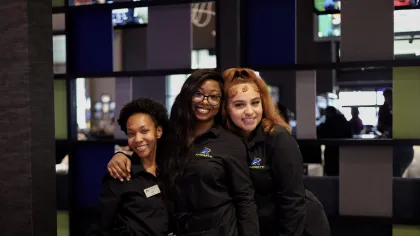Three smiling female employees in black uniforms posing together inside a modern establishment with colorful wall panels.