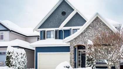 Snow-covered modern two-story house with stone facade and blue siding under cloudy sky in winter.