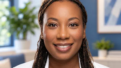 Smiling woman with long braided hair wearing white coat in bright modern office with plants and artwork.