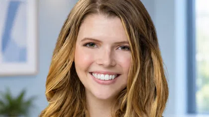 Smiling woman with long wavy blonde hair in a black blazer and teal top in a modern office setting