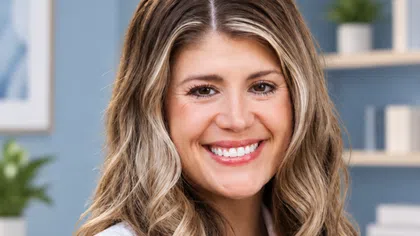 Smiling woman with long wavy hair wearing a white lab coat in a bright office setting