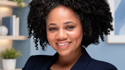 Confident young woman with curly hair smiling, wearing a navy blazer indoors with plants and books in background