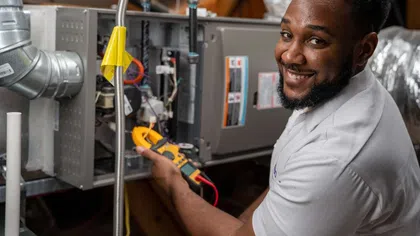 Technician in white shirt smiling while using a multimeter to service HVAC system in an attic.