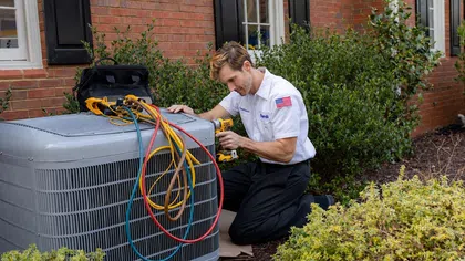 Technician in white uniform repairing outdoor air conditioning unit with colorful cables and power drill beside a brick house.