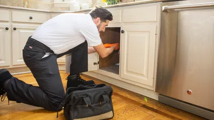 Plumber inspecting under kitchen sink, wearing orange gloves, with toolbox on wooden floor near dishwasher