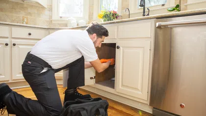 Plumber kneeling in a kitchen fixing pipes under the sink with tools and a black toolkit on wooden floor.