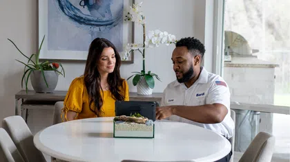 Technician explaining HVAC system details to a woman at a white dining table in a modern room with plants.