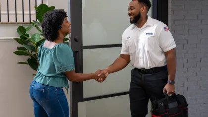 Technician in uniform shaking hands with woman at door carrying a black tool bag in a modern home.