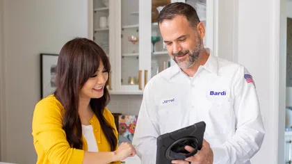 HVAC technician in white uniform shows tablet to smiling woman in yellow sweater in a modern kitchen