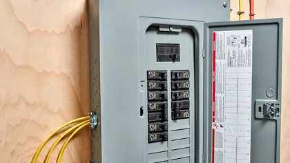 Close-up of a residential electrical panel with circuit breakers and yellow wires on a plywood background.