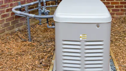 A grey generator installed outside a brick wall, surrounded by mulch and piping.