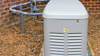 A grey generator installed outside a brick wall, surrounded by mulch and piping.