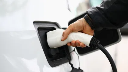 Person plugging a white electric vehicle charger into a white car at a charging station outdoors.